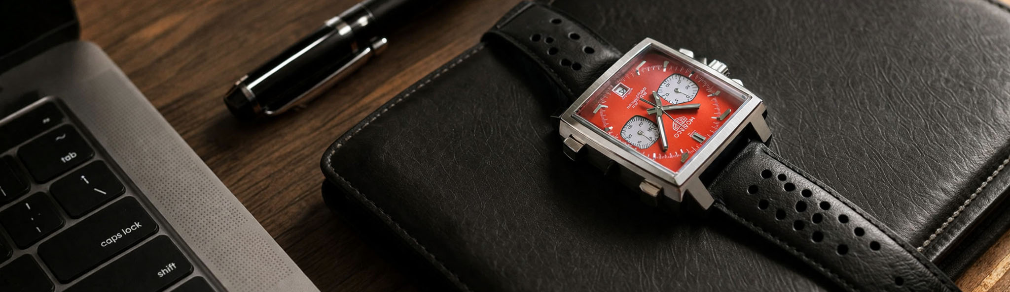 Red dial square chronograph watch with black leather strap placed on a leather notebook beside a laptop and pen on a wooden desk